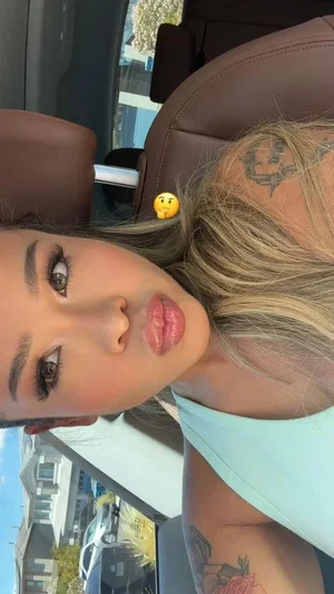 Woman with blonde hair and tattoos in car, wearing light blue top, making playful face.