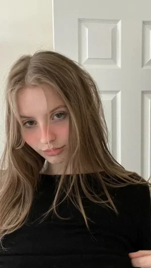 Woman with long blonde hair wearing black top, indoors near white door.