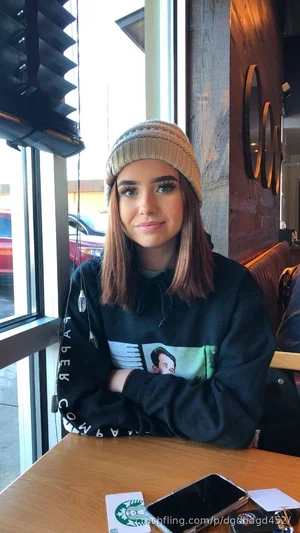 Woman with brown hair wearing beanie and black hoodie, sitting at table in cafe.