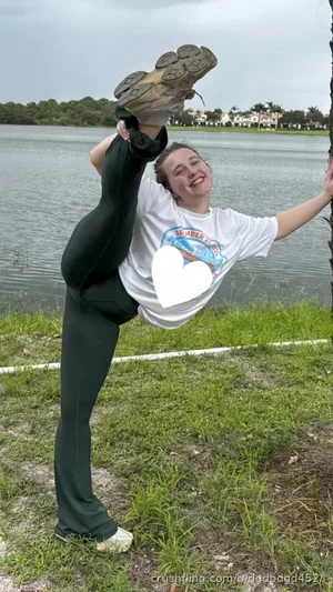 Woman in white t-shirt and black leggings doing a high kick by a lake, smiling.