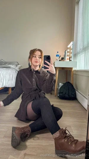 Woman in dark shirt and tights sitting on floor, taking mirror selfie in bedroom.