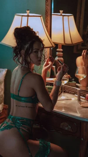 Woman in green lingerie sits at vanity, holding makeup brush, mirror reflection visible.