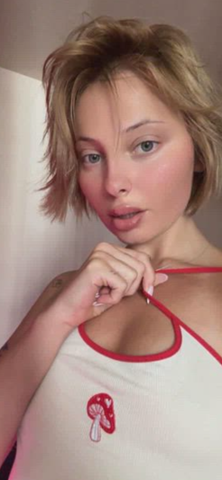 Woman with short blonde hair in white top with red trim and mushroom design, indoors.