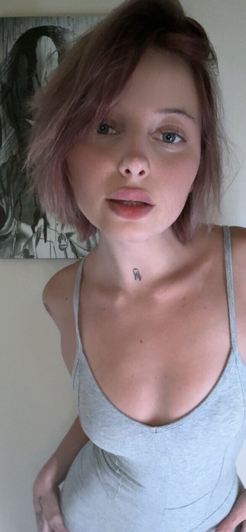 Woman with short pink hair in gray tank top, standing indoors near wall art.