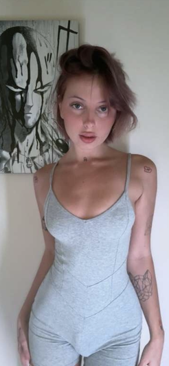Woman in gray tank top and matching pants, standing indoors with tattoos visible on arms and neck.