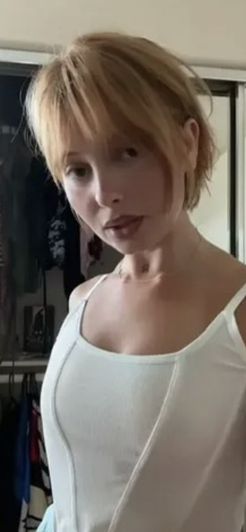 Woman with short blonde hair in white tank top, standing indoors near closet.