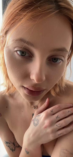 Woman with short red hair and tattoos, topless, hand on chest, indoor setting.