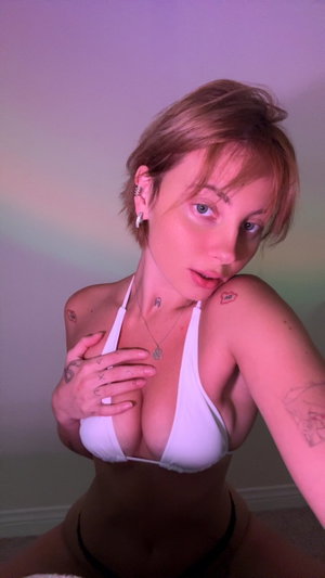 Woman with short blonde hair in white bikini top and black bottoms, posing indoors with tattoos
