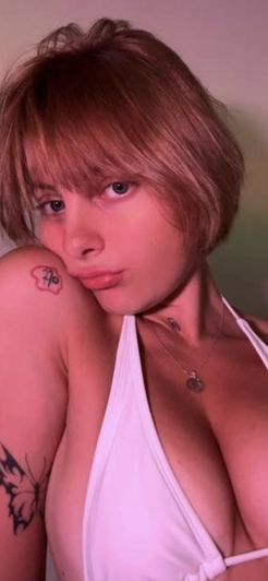 Woman with short brown hair and tattoos wearing white top, posing indoors.