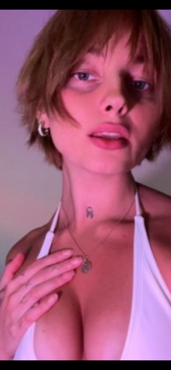 Woman with short hair in white top, hand on chest, indoor setting.