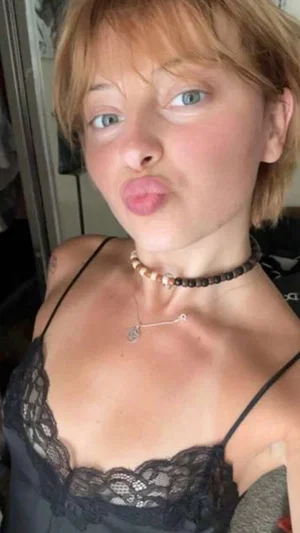 Woman with short blonde hair in black lace lingerie, making a playful face indoors.