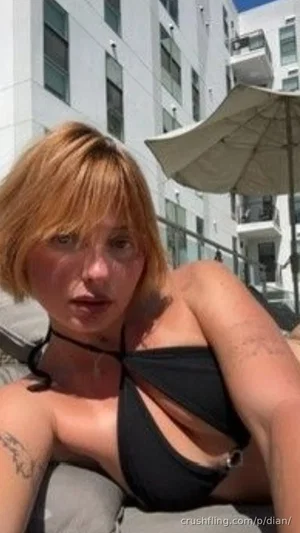 Woman with short blonde hair in black bikini top, sitting outdoors near buildings and umbrella.