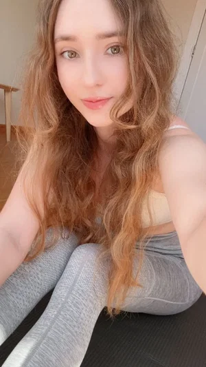 Woman with long wavy hair in light top and gray leggings sitting on mat indoors.