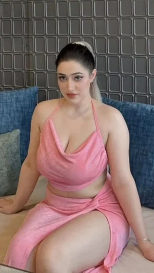 Woman in pink two-piece outfit sitting on bed with blue pillows and textured wall behind.