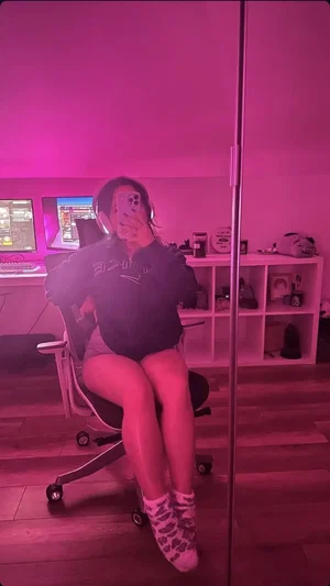 Woman in black hoodie and shorts sits in office chair, taking mirror selfie in pink-lit room.