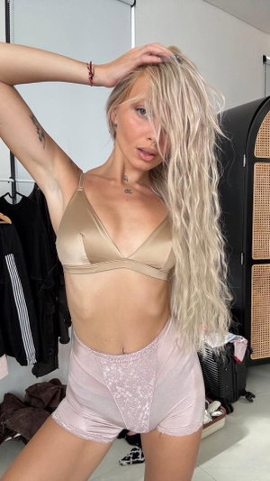 Blonde woman in beige bra and pink lace shorts posing indoors.