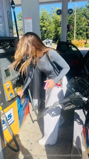 Woman in gray long-sleeve top and white pants at gas pump, holding phone.