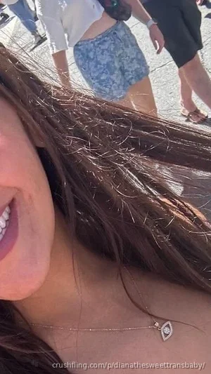Woman with long brown hair smiling, wearing a necklace, outdoors with others in background.