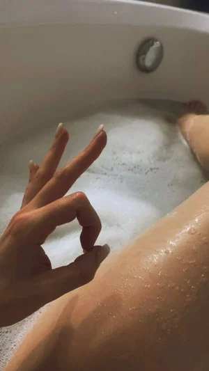 Woman with bare legs and hand in bubble bath, indoor setting.