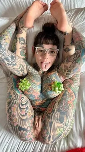 Tattooed woman with glasses lying on bed, legs raised, tongue out, green bows on chest.