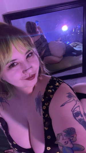 Woman with tattoos in black lace top and underwear, sitting indoors under purple light.