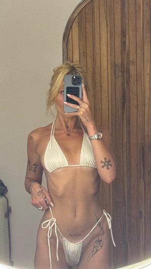 Blonde woman in white string bikini with tattoos in mirror selfie.