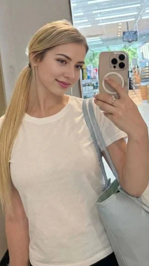 Blonde woman in white t-shirt takes mirror selfie in store, holding phone and bag.
