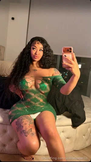 Woman with long curly hair in green sheer dress with cutouts, sitting on bed, taking mirror selfie.