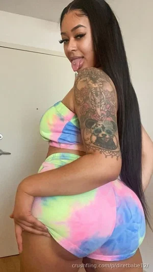Woman with long black hair and arm tattoo wears tie-dye crop top and shorts, posing indoors.