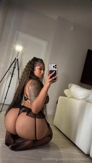 Woman with long curly hair in black lace lingerie and fishnet stockings, kneeling in a bedroom.