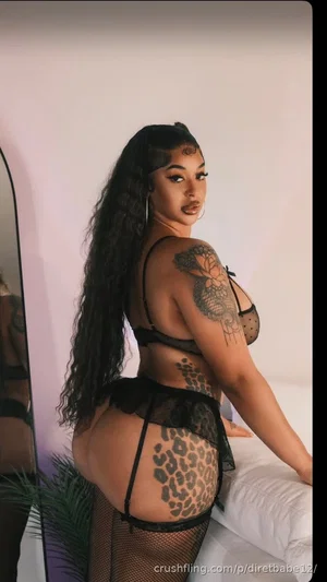Woman with long dark hair and tattoos wears black lace lingerie, posing by a bed indoors.