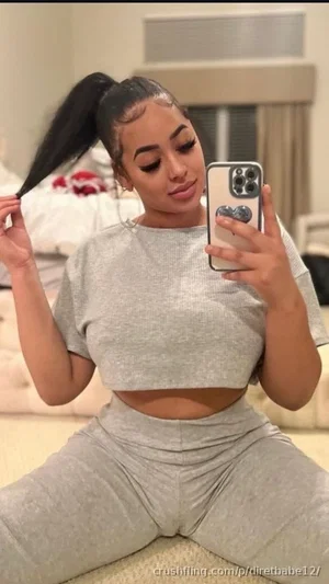 Woman in gray crop top and matching pants sits on floor, holding phone for mirror selfie in bedroom.
