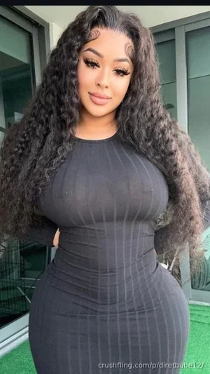 Woman with long curly hair in a form-fitting black dress, standing outdoors near glass doors.