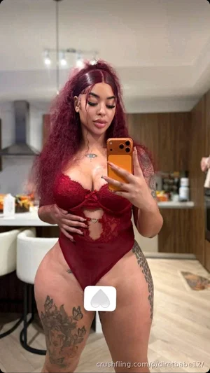 Woman with red hair and tattoos wears red lace bodysuit, taking mirror selfie in kitchen.