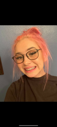 Woman with pink hair and glasses, wearing a dark t-shirt, smiling indoors.
