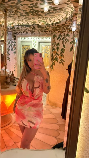 Woman with long hair in pink floral dress taking mirror selfie in bathroom.