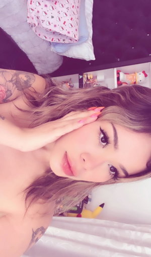 Woman with tattoos and long hair, topless, posing in bedroom with plush toys.