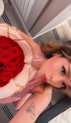 Woman in pink sparkly top holding red roses, posing indoors with tattoo on arm.