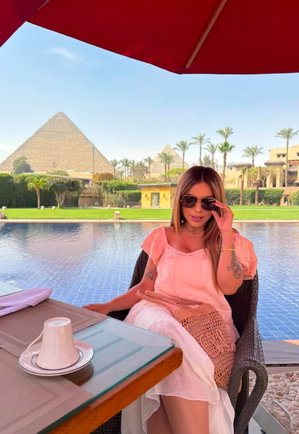 Woman in pink top and white skirt sits by pool under red umbrella, pyramids in background.