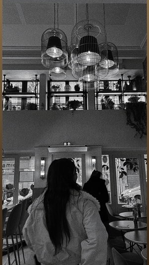 Woman with long dark hair in a fuzzy coat sits in a modern cafe with hanging lights and plants.