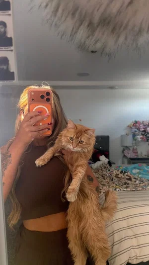Woman with tattoos and long hair in brown crop top holding cat in bedroom.