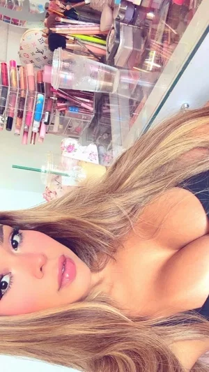 Woman with long blonde hair in black top, posing in front of makeup collection.
