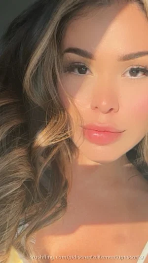 Woman with wavy brown hair, wearing a white top, in a close-up indoor selfie.