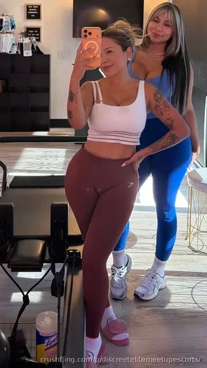 Two women in athletic wear pose for a mirror selfie in a gym; one wears a white crop top and brown