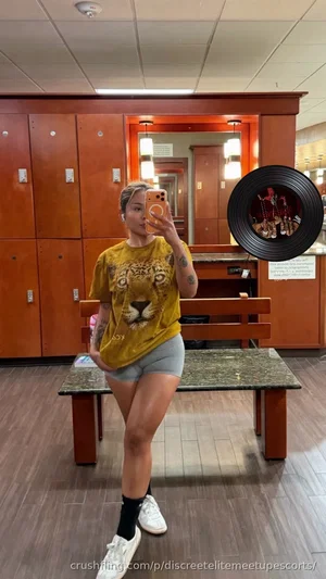 Woman in yellow tiger tee and gray shorts poses for mirror selfie in salon.