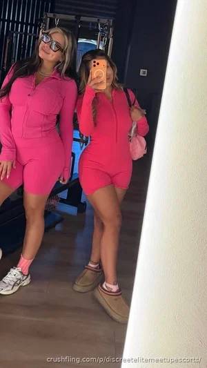 Two women in pink bodysuits pose for mirror selfie in indoor setting.
