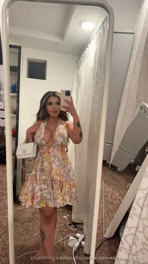 Woman in floral dress taking mirror selfie in bedroom, tattoos visible on arms and legs.