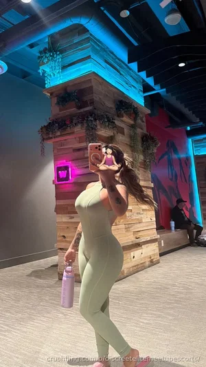 Woman in green bodysuit holding water bottle, standing in modern indoor space with wood paneling
