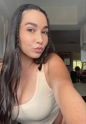 Woman with long dark hair in braids, wearing beige tank top, taking selfie indoors.