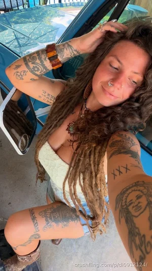 Woman with dreadlocks and tattoos wears a light top, posing near a blue vehicle outdoors.